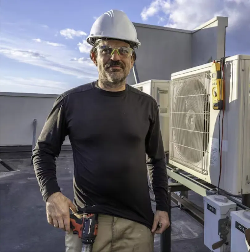 A man wearing safety equipment on a roof standing next to two HVAC units